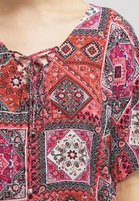 Top rouge et rose à motifs avec un décolleté en V agrémenté de lacets, des designs géométriques et des textures variées. Comprend des motifs floraux et en losange.