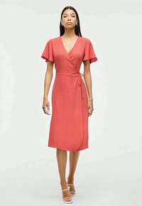 Vestido envelopado corais com mangas curtas e esvoaçantes, tecido texturizado, faixa na cintura, e comprimento até o joelho. Usado com sandálias brancas com tiras.