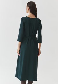 Robe vert foncé, mi-mollet, avec des manches trois-quarts, un col rond et une ceinture à la taille, fabriquée en tissu lisse et texturé.