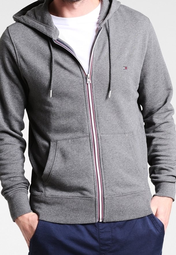 Homme portant un sweat à capuche gris zippé avec poches avant et cordons, mains dans les poches, par-dessus un T-shirt blanc et un pantalon bleu marine.