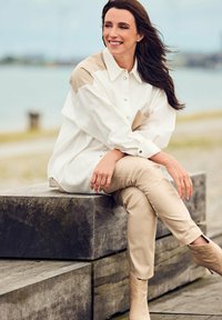 Witte oversized shirt met beige accenten, gecombineerd met strakzittende beige broek en nude enkellaarsjes, zittend op houten banken aan de waterkant.