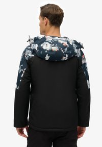 Herenjack in zwart met kap, met een kleurrijk, gestructureerd patroon op schouders en mouwen, benadrukt door een gladde zwarte onderkant.