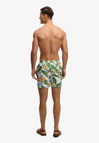 Homme debout pieds nus, de dos, portant un short de bain à fleurs avec des feuilles vertes et des fleurs orange sur fond blanc.