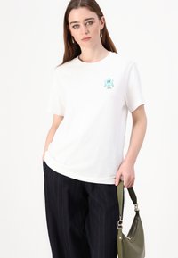White cotton t-shirt featuring a light blue cartoon character on the left chest. Styled with loose black pinstripe trousers and a green handbag.