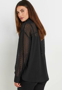 Blusa nera trasparente con motivo a spina di pesce, maniche lunghe e colletto, con una vestibilità morbida e un orlo posteriore leggermente più lungo.