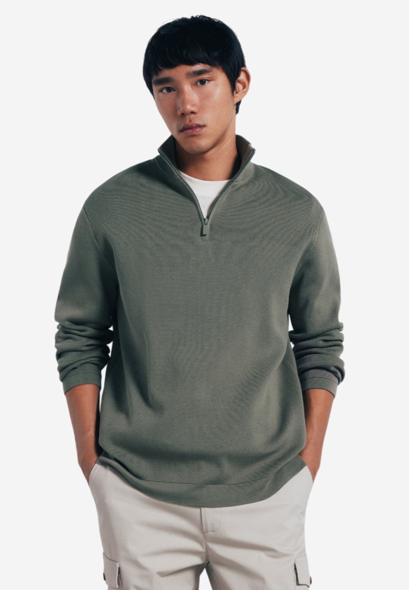 Maglione verde oliva, tessuto a trama, con colletto alto, chiusura a mezza zip e polsini a costine, indossato sopra una camicia bianca.