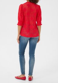 Top in pizzo rosso con maniche a palloncino, abbinato a jeans skinny blu e ballerine rosse, che mostrano motivi floreali e una finitura testurizzata.