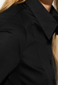 Svart skjorta med knappar och spetsig krage, tillverkad av mjukt tyg. Har en figurnära silhuett och subtila sömnadsdetaljer på axlarna.