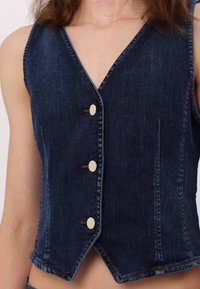 Džínová vesta v tmavě modré barvě s výstřihem do V, předními knoflíky a pečlivě prošívanými švy. Vesta má zkrácený design a mírně rozšířený lem.