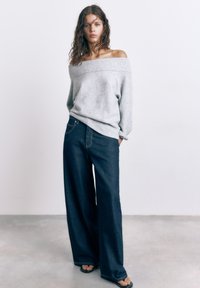 Mladá žena s vlnitými vlasy, která nosí šedý svetr s odhalenými rameny, tmavě modré široké džíny a sandály v jednoduchém vnitřním prostředí.