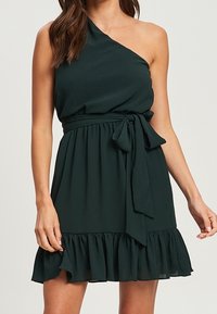 Sukienka na jedno ramię w ciemnozielonym kolorze, uszyta z lekkiego materiału, z dopasowaną talią i falbaniastym dołem. W komplecie jest pasujący pasek do wiązania.
