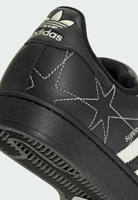 Black synthetic sneaker with white accent, featuring star pattern stitching, textured sole, and classic three-stripe design.