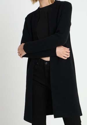 Femme portant un long cardigan noir ouvert sur un haut noir et un pantalon noir, les bras croisés à la taille.