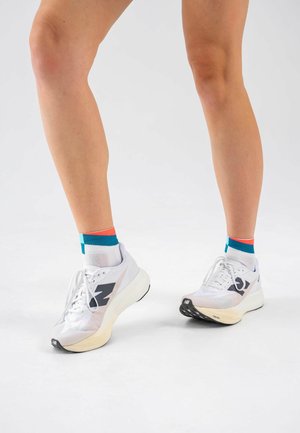 Beine, die weiße Laufschuhe mit schwarzen Logos und dicken Sohlen tragen, kombiniert mit weißen Socken mit blauen und roten Streifen.