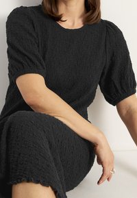 Černé strukturované šaty s krátkými puff rukávy a vlnitým lemem. Tkanina má zmačkaný vzhled, což zvyšuje vizuální zajímavost.