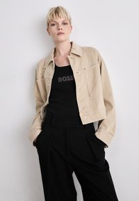 Giacca in denim beige con design corto, caratterizzata da chiusura a bottoni e dettagli sulle tasche, indossata sopra una canotta nera con la scritta luccicante "BOSS".