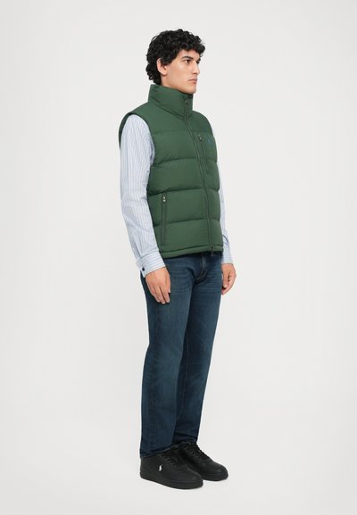 Hombre con chaleco acolchado verde sobre camisa de rayas, jeans azules y zapatillas negras de pie contra un fondo blanco liso.