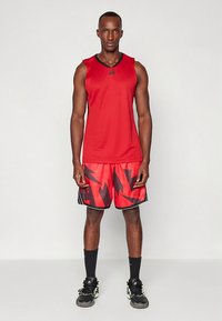 Rdeč športni top z črnimi poudarki, kombiniran z rdečimi kratkimi hlačami z črnim cikcak vzorcem. Oblečeno s črnimi športnimi copati.