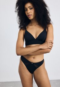 Zwarte bralette met een V-halsontwerp en dunne bandjes, gecombineerd met een bijpassende zwarte string met meshaccenten. Zachte, gladde stofstructuur.