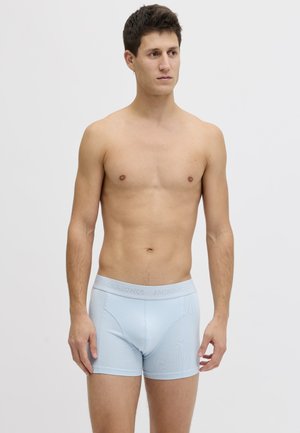 Homme debout face à l’avant, portant un boxer bleu clair avec une ceinture visible, sur un fond uni clair.