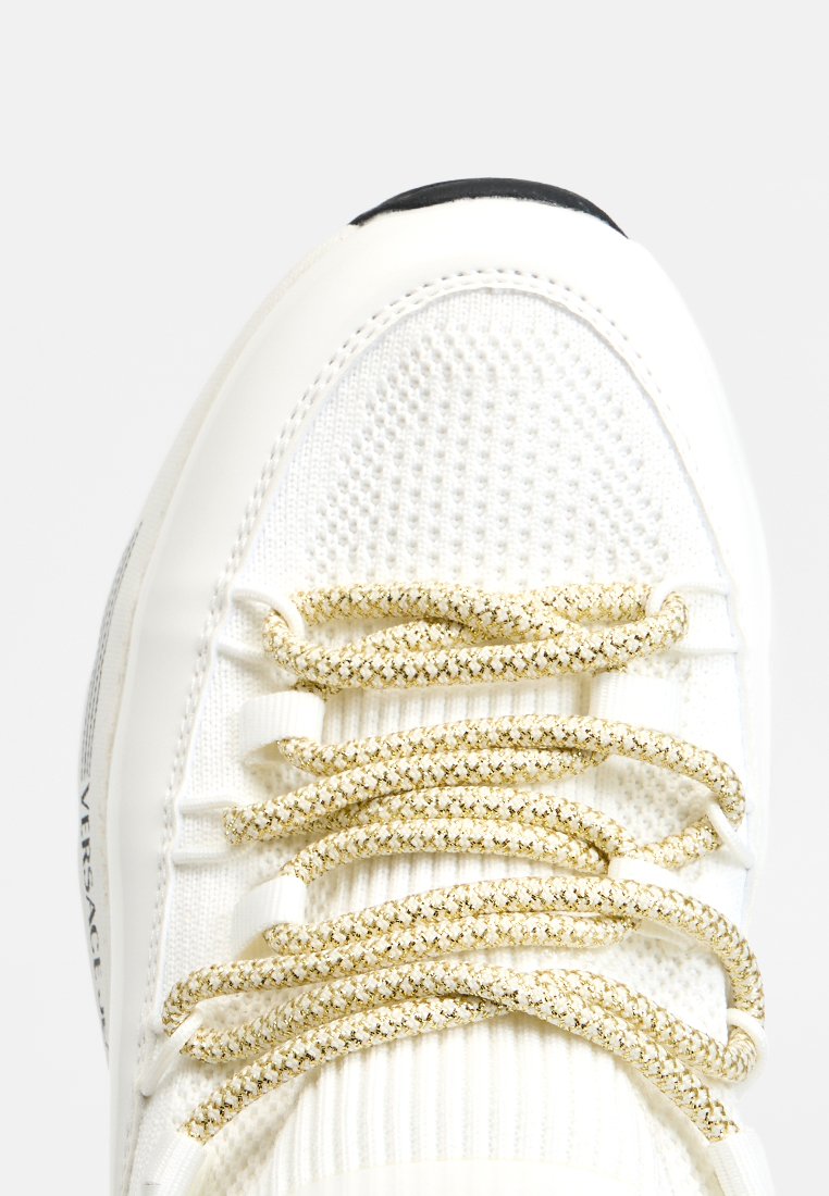 Zapatillas de punto blancas con cordones redondos acentuados en dorado, enhebrados a través de lazos de tela, que presentan una punta negra y una marca parcial en el costado.