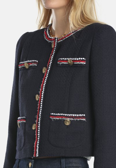 Veste bleu marine avec tissu texturé, boutons dorés et bordure rouge et blanche sur les poches et les coutures ; présente un design court et structuré.
