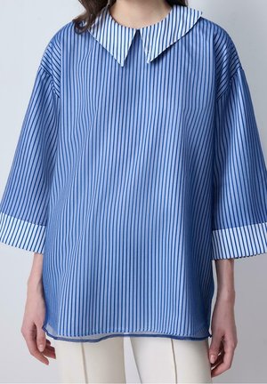 Femme portant un chemisier bleu ample à fines rayures verticales, un col blanc pointu et des manches trois-quarts avec des poignets blancs.