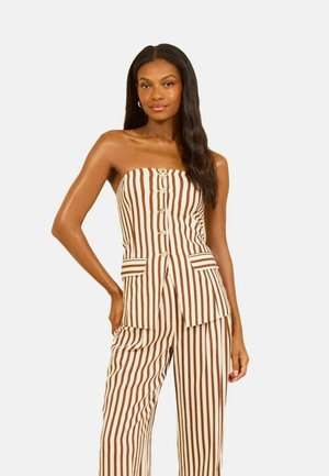 Vrouw draagt een strapless bruine en witte jumpsuit met verticale strepen, knopen en voorzakken met kleppen, staand met één hand op de heup.