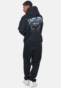 Sort hættetrøje med blå grafisk tekst "LIMITLESS DROPSIZE" på ryggen, med hvide lynakcenter. Sammen med sorte bukser og hvide sko.