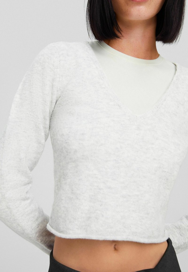 Frau in einem hellgrauen, flauschigen, cropped V-Ausschnitt-Pullover über einem weißen Oberteil mit schwarzen Hosen, stehend vor einem schlichten grauen Hintergrund.