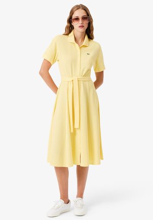 Femme portant une robe midi boutonnée à manches courtes de couleur jaune pâle avec une ceinture à la taille, des baskets blanches et des lunettes de soleil marron, debout avec les mains dans le dos.