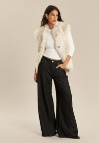 Faux fur vest in crême met bruine knopen, gecombineerd met hoge taille zwarte brede jeans. Gladde textuur en een relaxte pasvorm.
