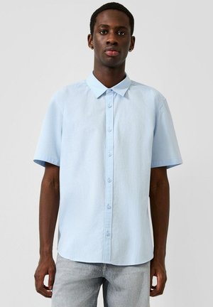 Mann trägt hellblaues kurzärmeliges Hemd mit Knöpfen und hellgraue Jeans, steht vor einfachem Hintergrund.