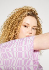 Blusa roxa com padrão apresentando designs geométricos ousados em branco. Modelo tem cabelo loiro encaracolado, parcialmente cobrindo o rosto.