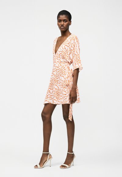 Γυναίκα που στέκεται φορώντας ένα κοντό, λευκό φόρεμα τύπου wrap με πορτοκαλί αφηρημένα μοτίβα και λευκά πέδιλα ψηλοτάκουνα με λουράκι στον αστράγαλο, ουδέτερο φόντο.