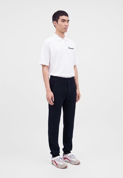 Homme debout portant un polo blanc, un pantalon noir et des baskets claires avec des accents rouges sur un fond uni.