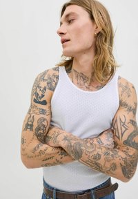 Balta juostuota maikutė, demonstruojanti sudėtingus tatuiruotes ant rankų, dėvima su šviesiai mėlynomis džinsomis ir ruduoju diržu. Natūralūs plaukai krinta žemiau pečių.