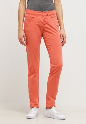 Pantalon corail ajusté avec boutonnière boutonnée, poches avant et arrière, porté avec un haut gris et des baskets blanches sur un fond uni.