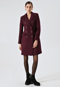 Cappotto bordeaux doppiopetto con bottoni circolari in argento, forma sartoriale, due tasche anteriori e tessuto texturizzato. Stivaletti neri alla caviglia con zip.