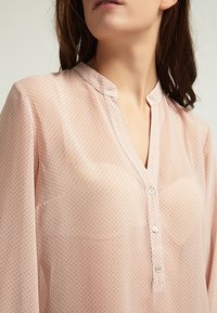 Blusa rosa chiaro con un sottile motivo geometrico, colletto alla mandarina e pattina con bottoni. Tessuto leggermente trasparente con maniche lunghe.