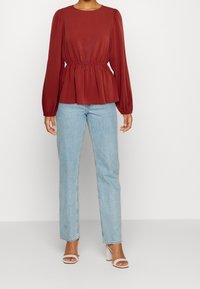 Červená peplum blůza s dlouhými rukávy a elastickým pasem, kombinovaná se světlomodrými džíny rovného střihu a bílými sandály na podpatku.