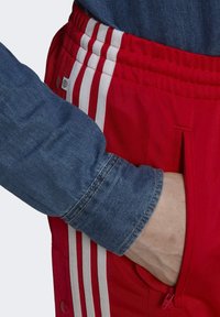 Czerwone spodnie sportowe z białymi paskami i zamykaną na zamek kieszenią. Jasnoniebieska koszula dżinsowa z długim rękawem, na którym widoczny jest mankiet przy nadgarstku.