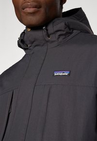 Čierna nepremokavá bunda s vysokým golierom, kapucňou na stiahnutie a zlatými akcentami. Obsahuje logo značky Patagonia a predné vrecká.