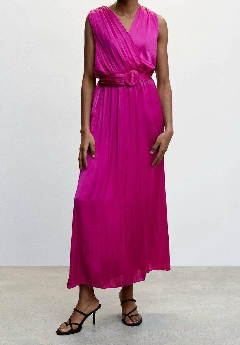 Femme portant une robe longue plissée rose vif sans manches, ceinturée à la taille, avec des sandales noires à talons hauts à brides, debout sur un sol uni.