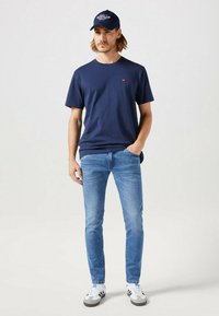 Marineblauwe katoenen t-shirt met korte mouwen en een klein rood logo, gecombineerd met lichtblauwe jeans en witte sneakers met zwarte accenten, afgewerkt met een marineblauw pet.
