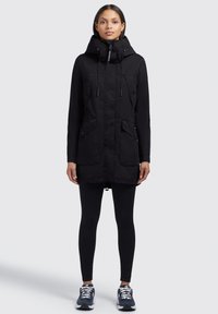 Parka noire avec capuche doublée, deux grandes poches avant et cordons de serrage réglables. Tissu lisse et design mi-long.