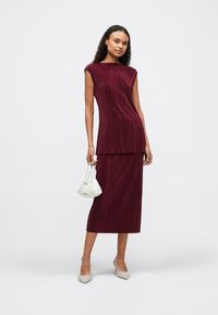 Βουργουνδίνο σετ δύο κομματιών, που περιλαμβάνει μια αμάνικη μπλούζα με κάθετη υφή και μια μίντι φούστα, συμπληρωμένο με μια λευκή τσάντα και μπεζ τακούνια.