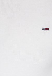 Valkoinen puuvillapaita, jossa on ribbikuvio. Paidassa on pieni brodeerattu logo punaisena, valkoisena ja sinisenä vasemmassa rintamuksessa.