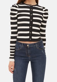 Žena oblečená v černo-bílém pruhovaném krátkém svetříku s perlovými knoflíky a vycpanými rameny, doplněná tmavě modrými džíny.