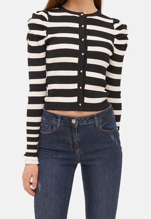 Mujer con un cárdigan corto de rayas horizontales en blanco y negro, con botones de perlas y mangas abullonadas, combinado con unos jeans de color azul oscuro.